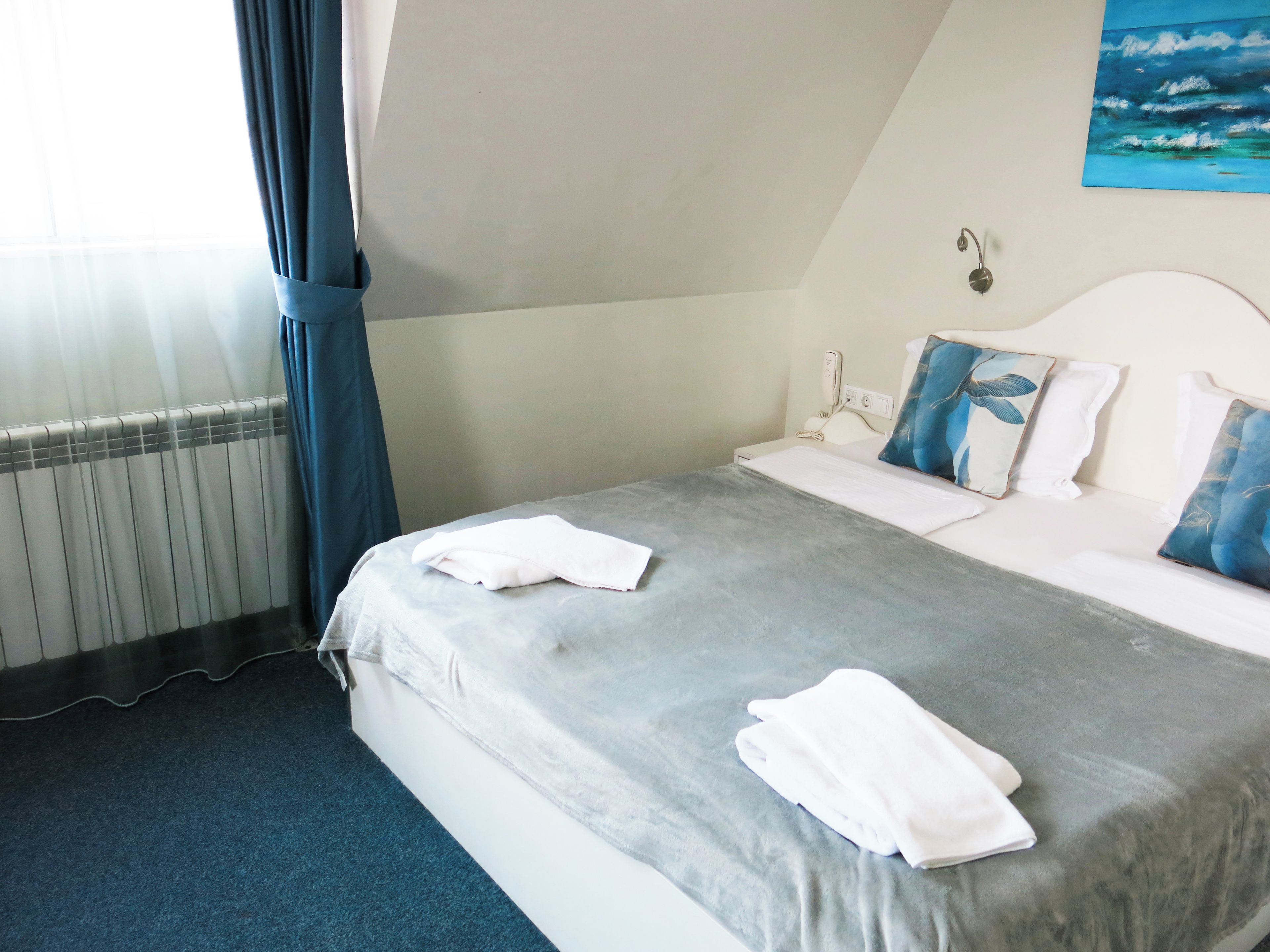 A hotel room with a bed, a bedside table, a nightstand, a wall painting, and a window with curtains.