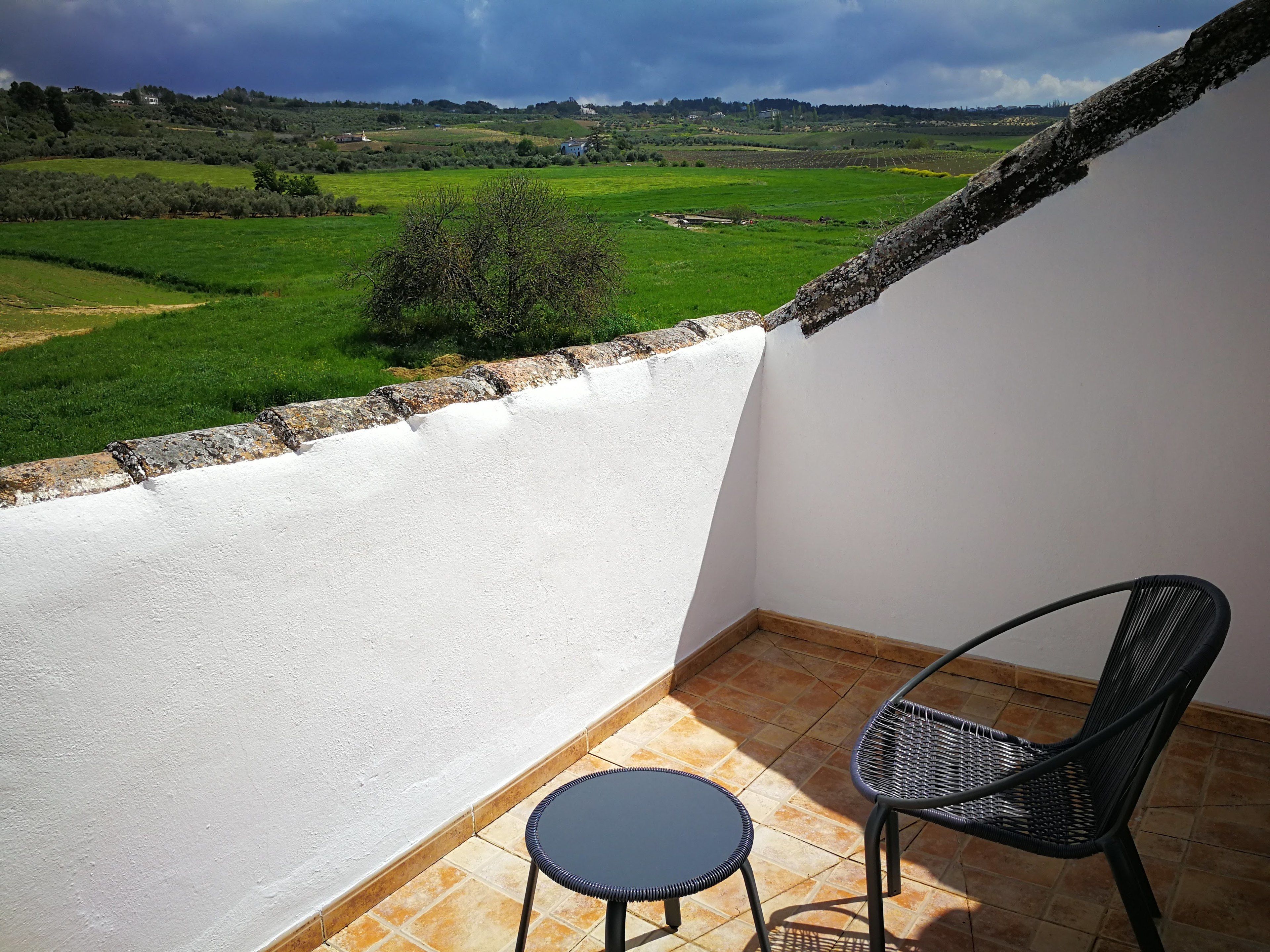 A balcony with a view of green fields and distant houses, a small round table, and a black wicker chair.