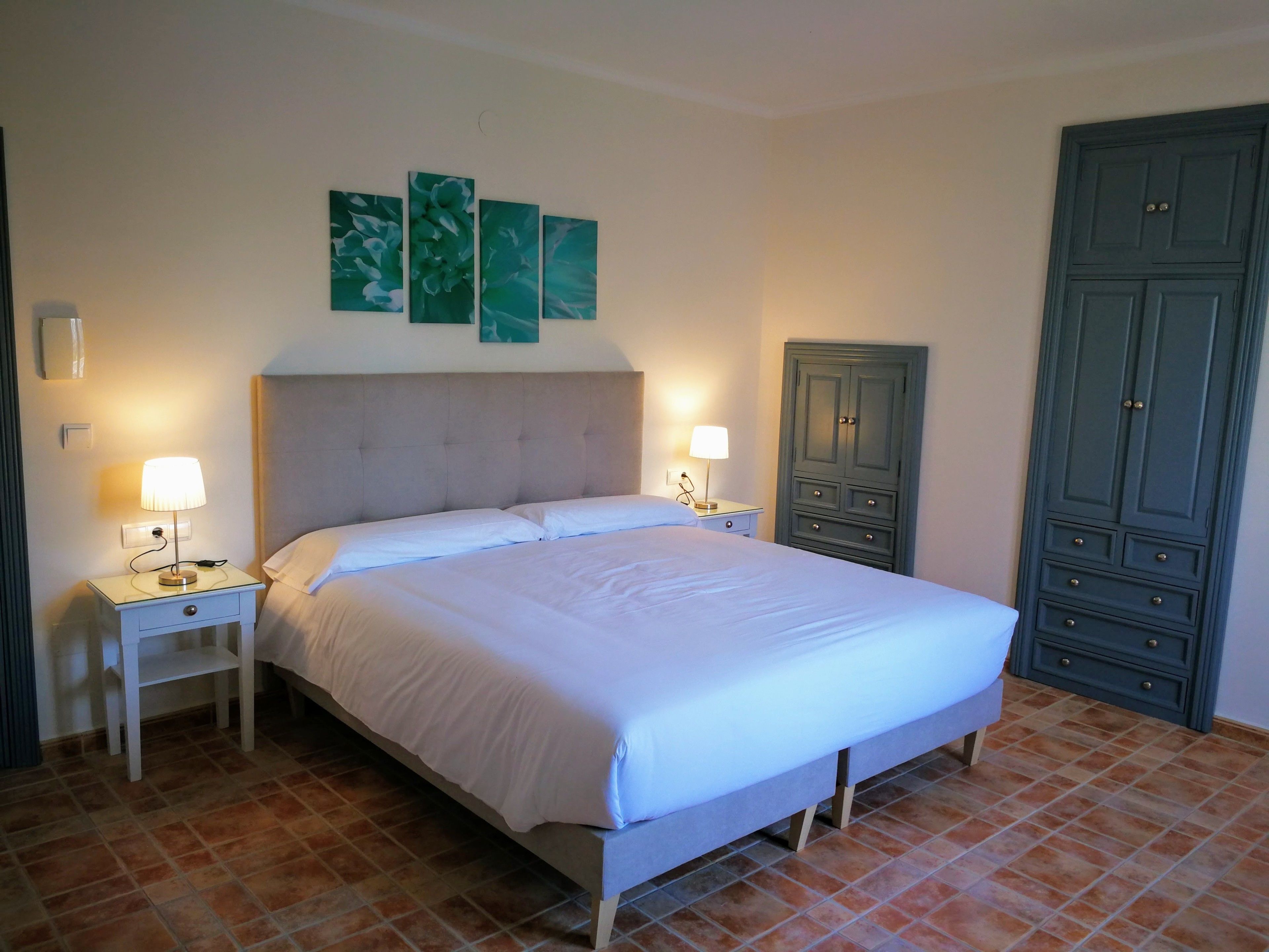 A neatly made bed with white linens, two bedside tables with lamps, a large wardrobe, and three wall-mounted paintings.