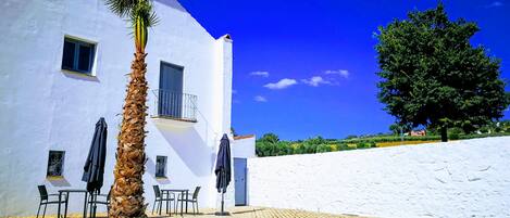 Un edificio blanco con un balcón, una palmera y un patio empedrado con sillas y mesas.