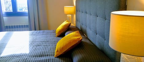 Una cama bien hecha con cabecera capitonada, dos almohadas y una lámpara de noche.