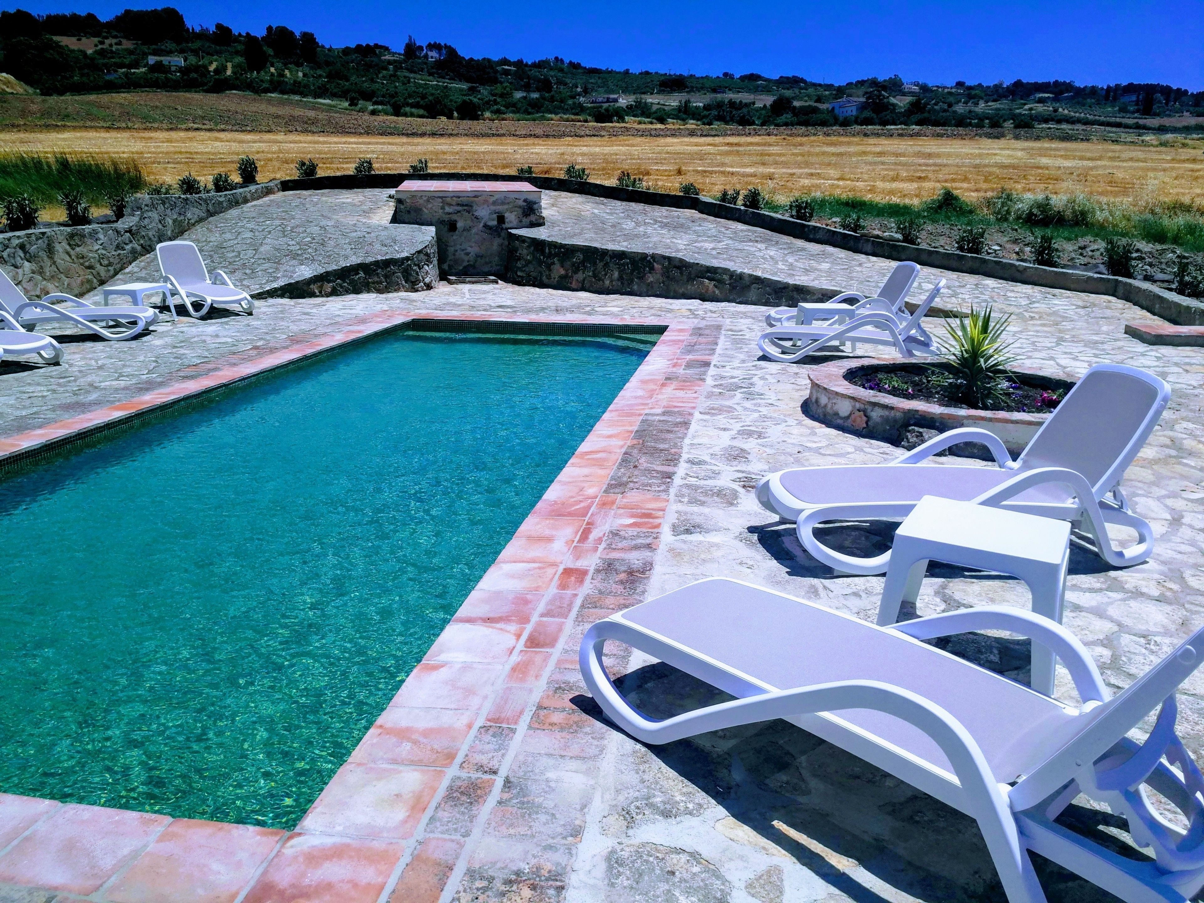 Una piscina rectangular con agua cristalina y azul, rodeada de baldosas de piedra y sillones blancos.