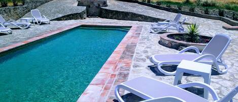 Una piscina rectangular con agua cristalina y azul, rodeada de baldosas de piedra y sillones blancos.