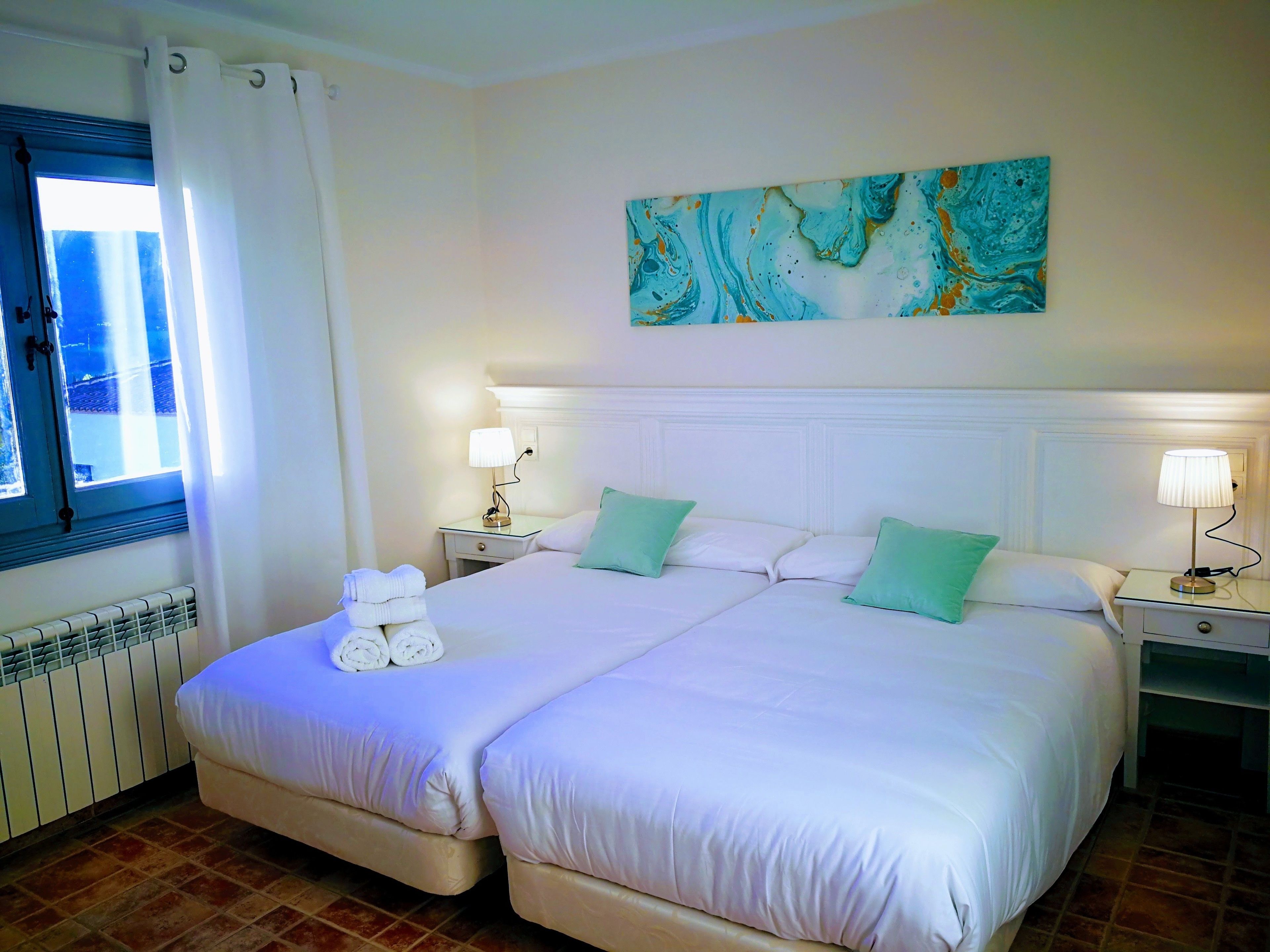 A double bed with white linens, two bedside tables with lamps, a radiator, and a window with curtains.