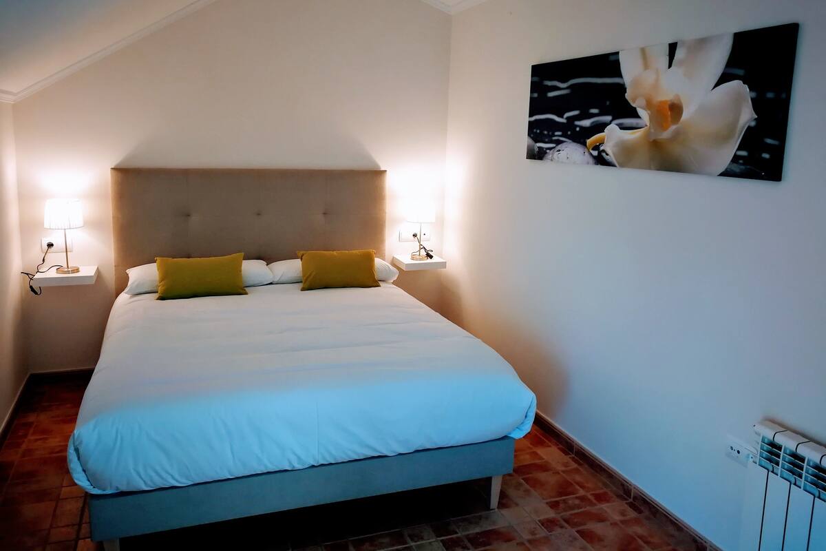 A neatly made bed with white and mustard pillows, two bedside lamps, and a wall-mounted picture of a bird.