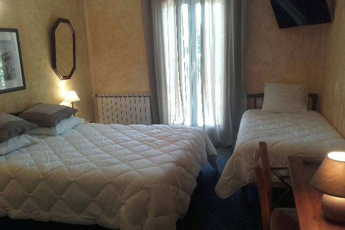 A hotel room with two beds, a radiator, a window with curtains, a TV on the wall, and a table with a lamp.