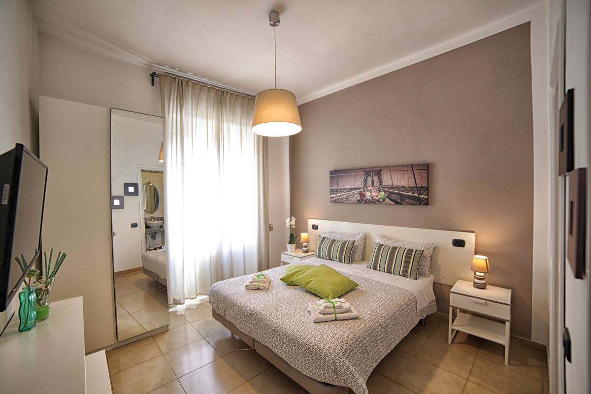 Ein Hotelzimmer mit einem großen Bett, einem Nachttisch mit Lampe, einem Spiegel, einem Bild an der Wand und einem durchgegangenen Tür erkennbaren Badezimmer.