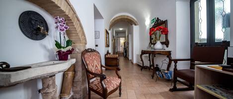 A hallway with a stone archway, a wooden chair, a table with red flowers, and a bookshelf.
