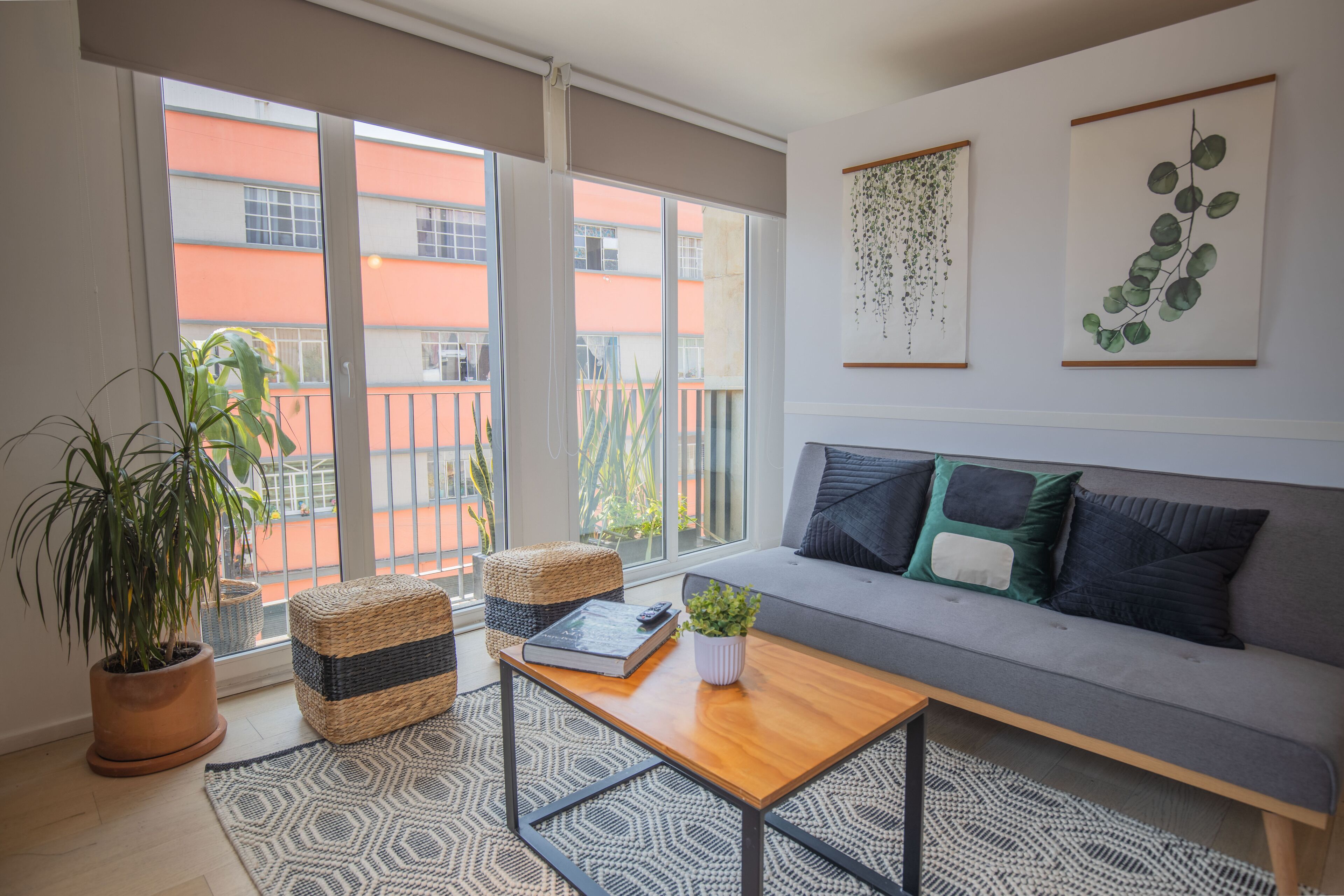 Un salon moderne avec un canapé gris, une table basse en bois, une plante en pot et une vue sur un immeuble orange, visible à travers de grandes fenêtres.