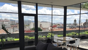 Un salon moderne avec un canapé d’angle foncé, deux petites tables rondes et une vue sur le paysage urbain à travers de grandes fenêtres.