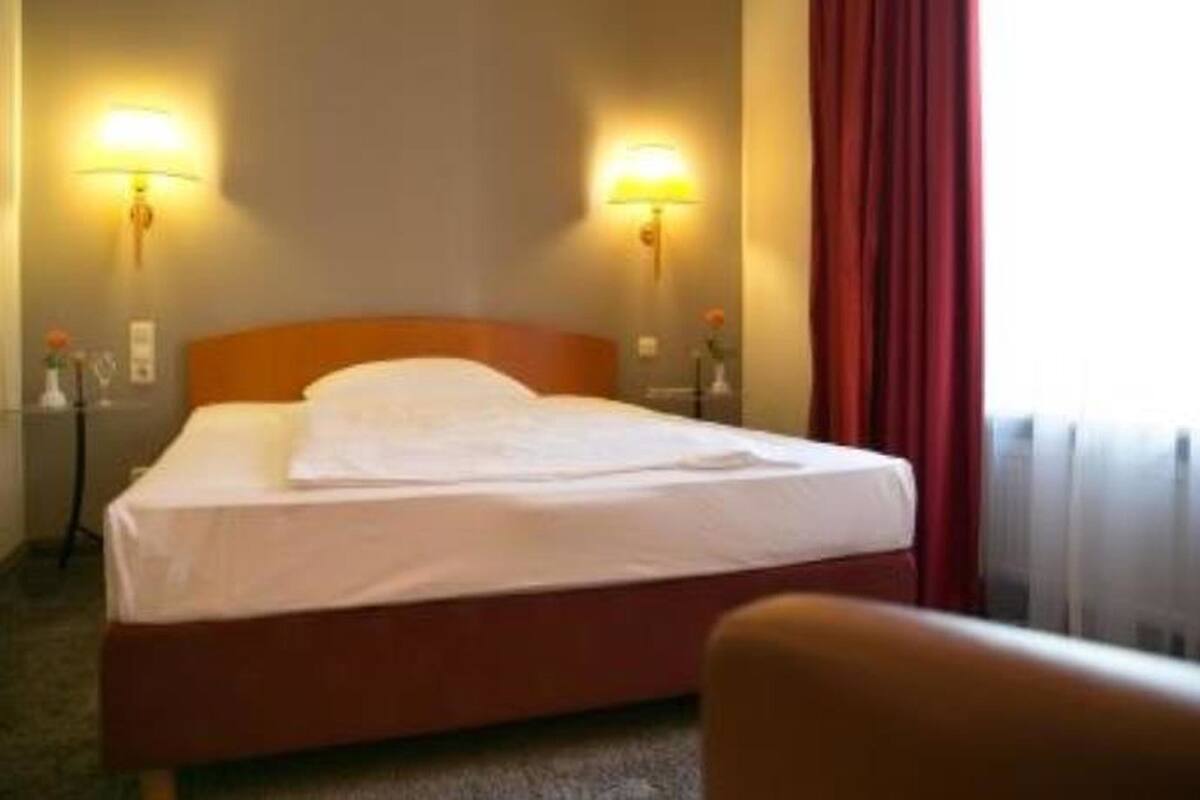 A hotel room with a bed, two wall-mounted lamps, a red curtain, and a chair.