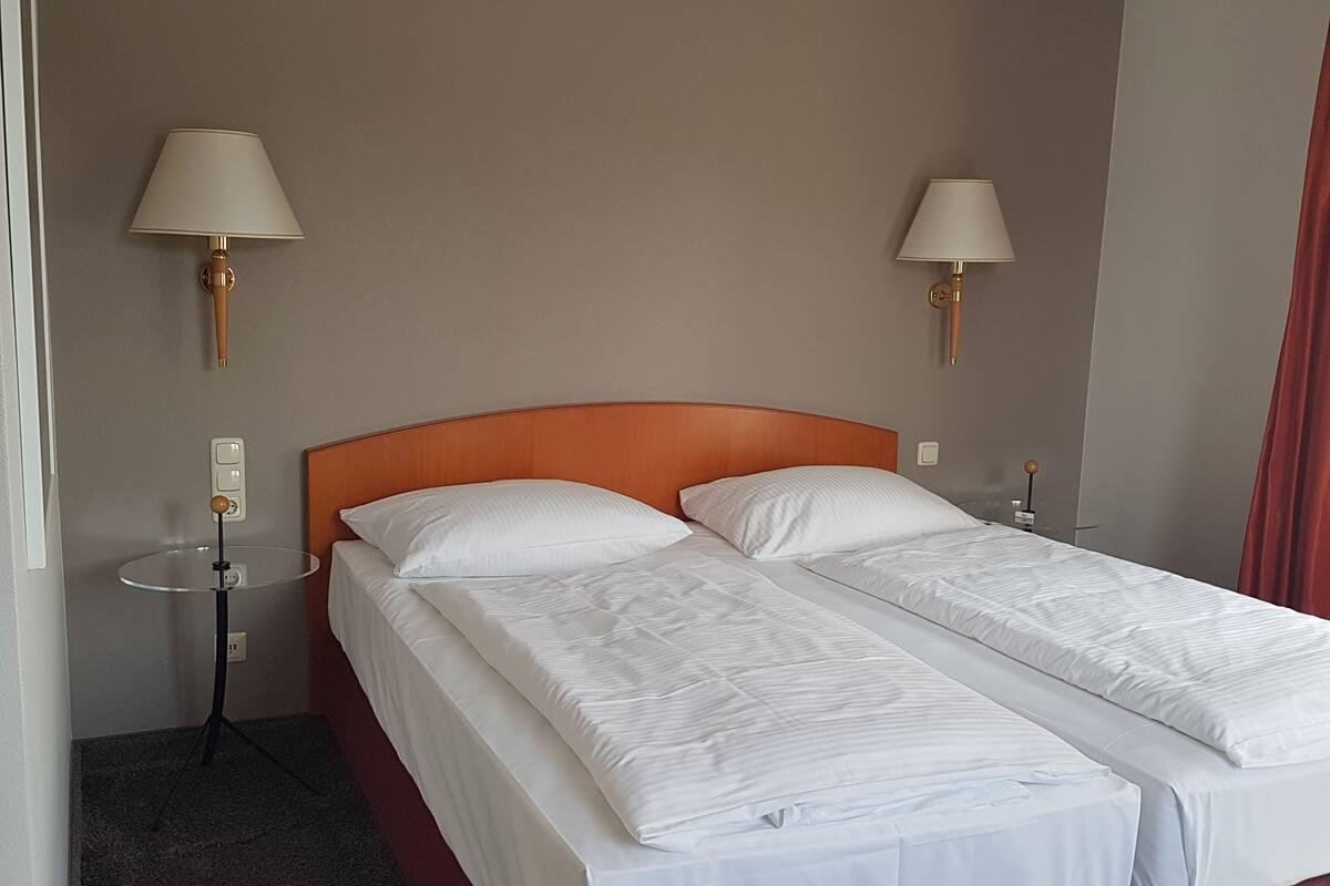 A hotel room with a double bed, two bedside lamps, a small table, and a red curtain.