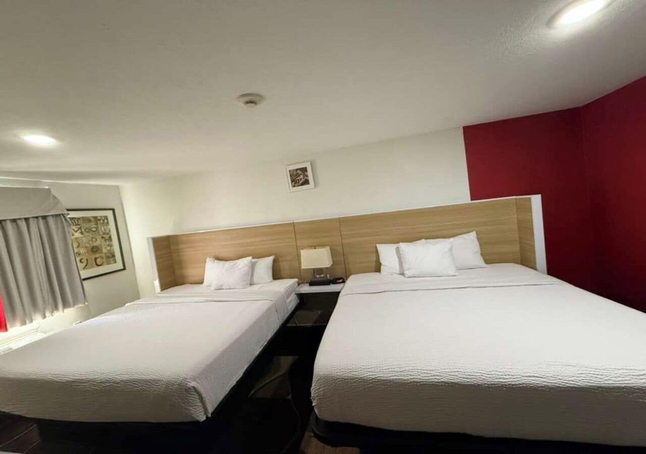 Habitación de hotel con dos camas, cabecero de madera, mesita de noche con lámpara y ventana con cortinas.