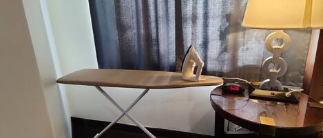 Una habitación compacta con una tabla de planchar plegable, un reloj digital y una lámpara sobre una mesita.