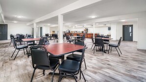 A spacious indoor area with multiple tables and chairs, a kitchen area in the background, and a wooden floor.