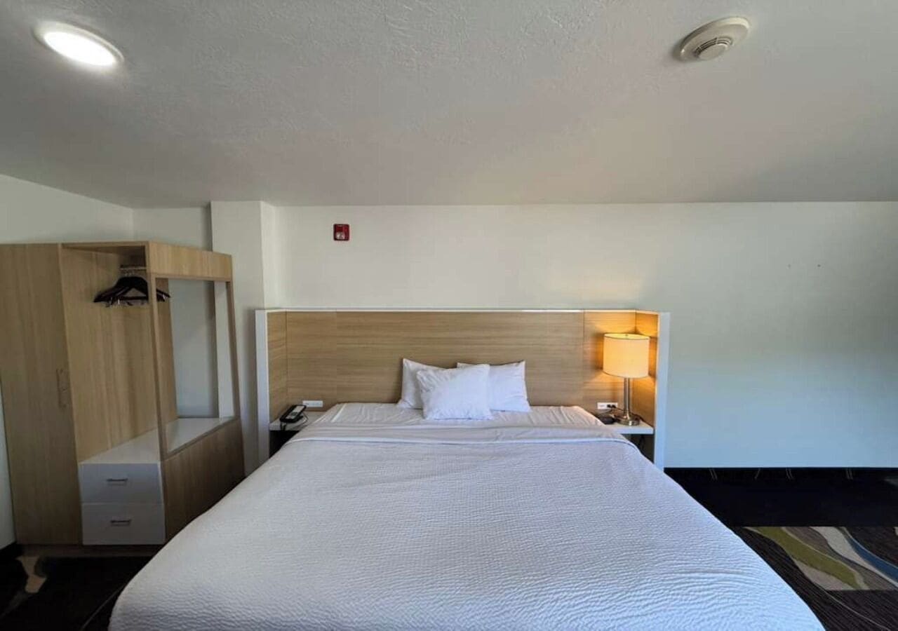 A hotel room with a large bed, a wooden wardrobe, a bedside table with a lamp, and a small desk.