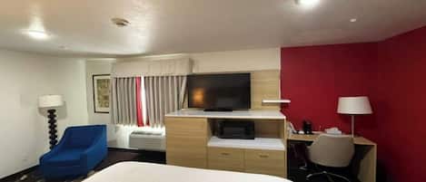 A hotel room with a large bed, a desk with a flat-screen TV, a blue chair, a desk lamp, and a red accent wall.