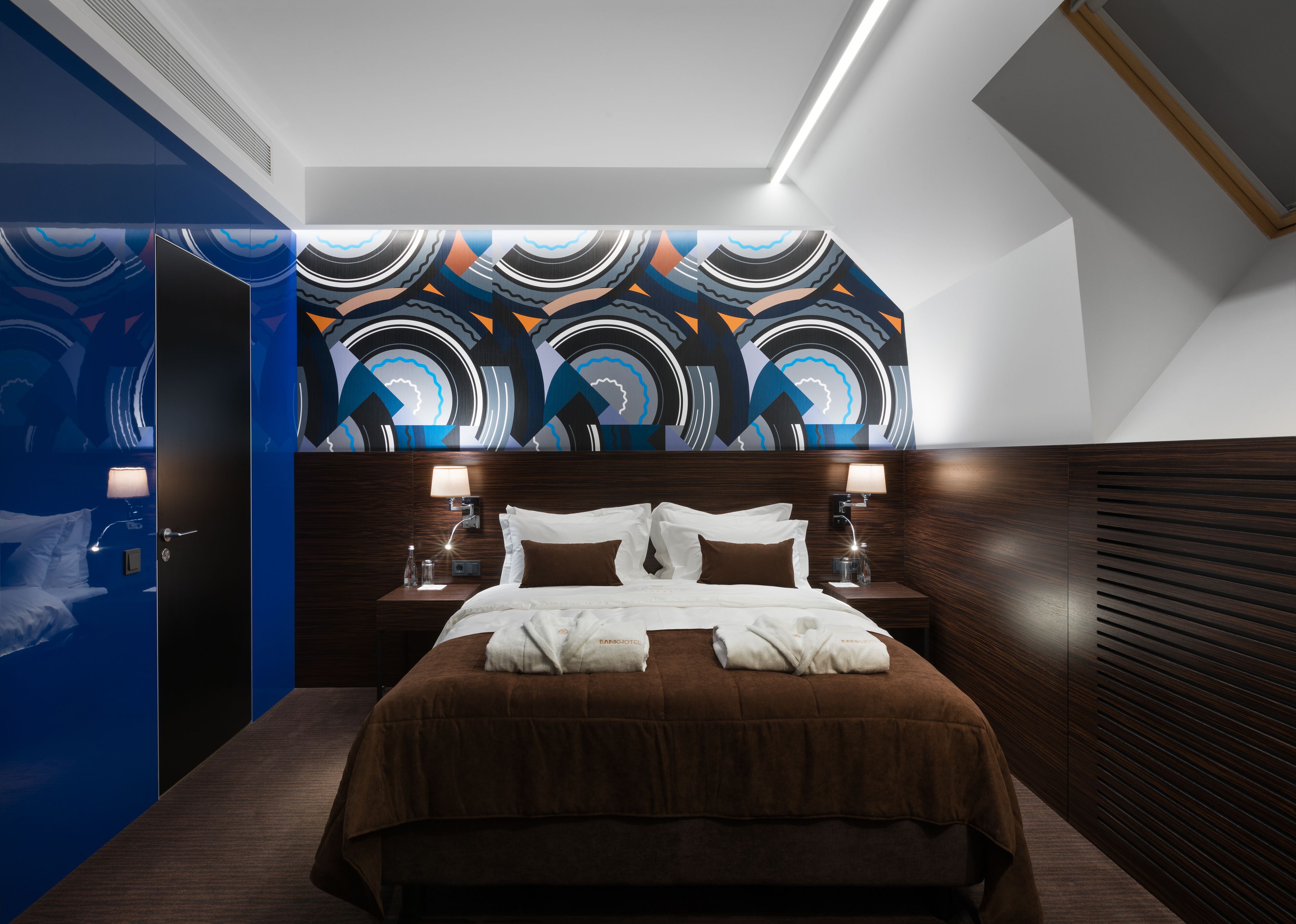 A modern hotel room with a large bed, wooden headboard, and a vibrant abstract wall mural.