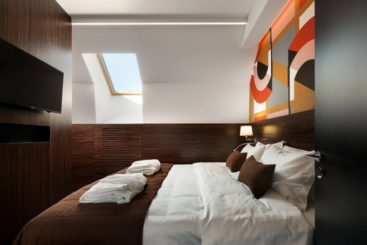 A modern hotel room with a large bed, a flat-screen TV, a skylight, and a wall-mounted lamp.