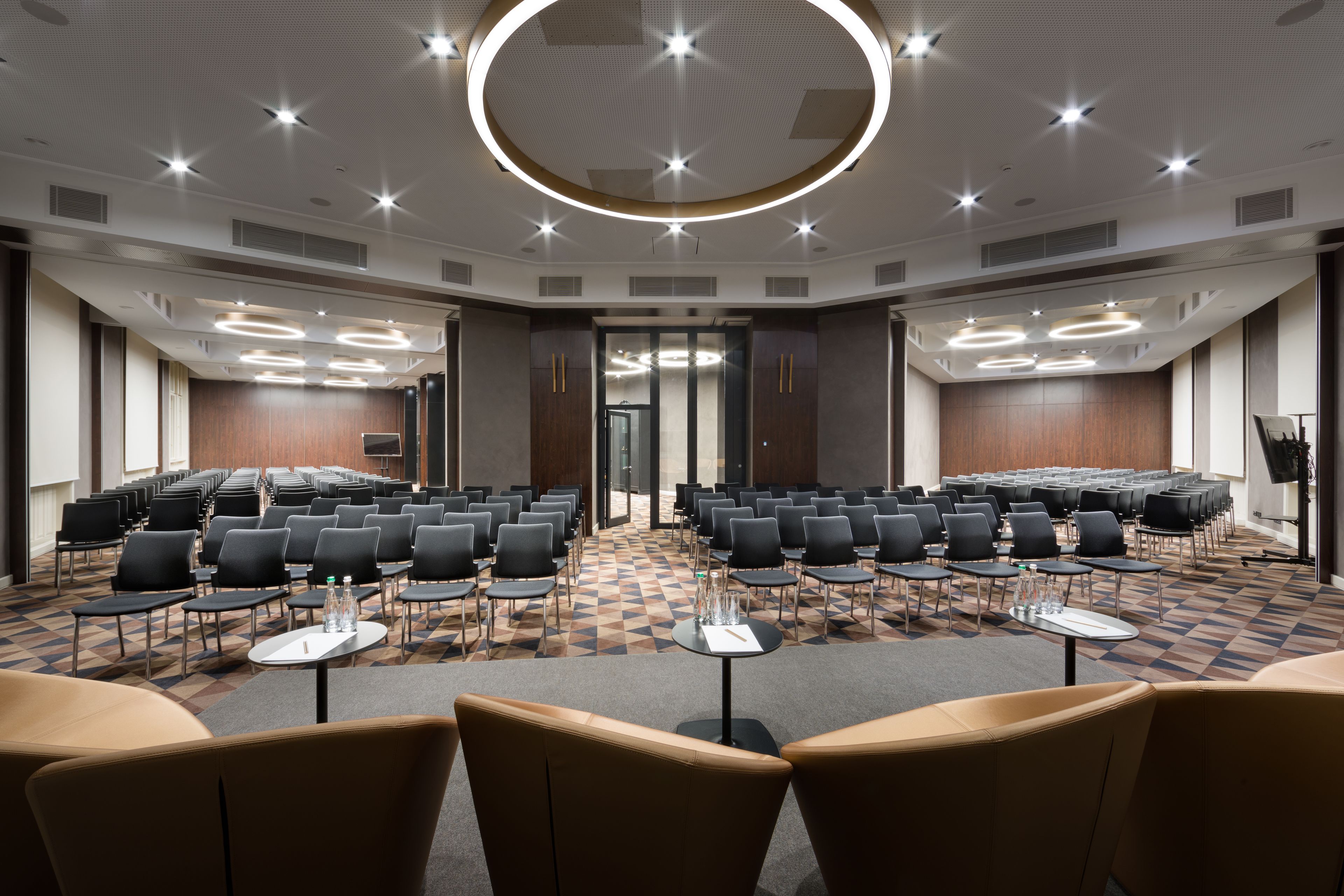 Uma sala de conferências com fileiras de cadeiras, mesas e um candeeiro circular no teto.