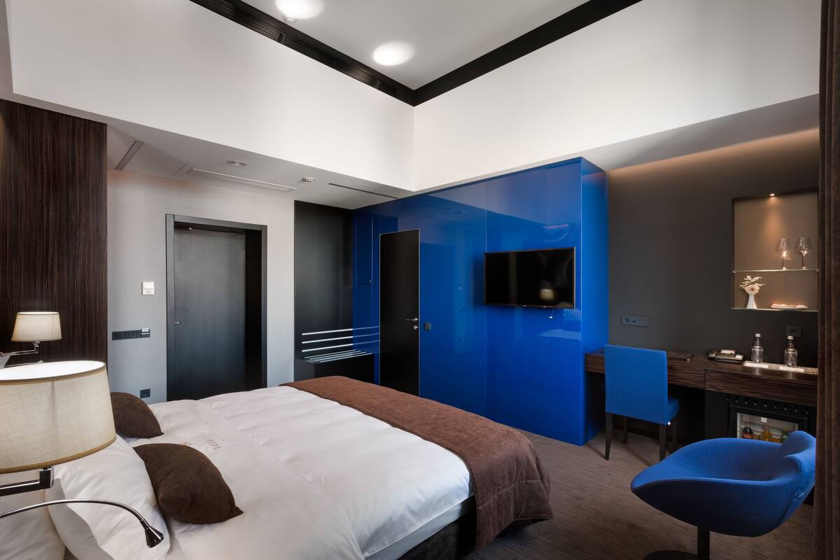 A modern hotel room with a large bed, a desk, a chair, a TV, and a minibar.