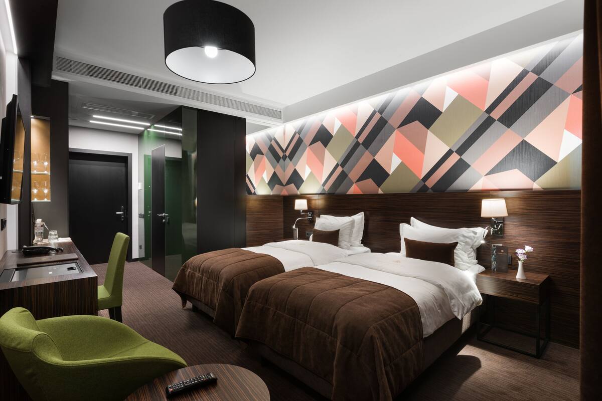 A hotel room with a large bed, two bedside tables, a desk with a chair, a green armchair, and a modern ceiling light.