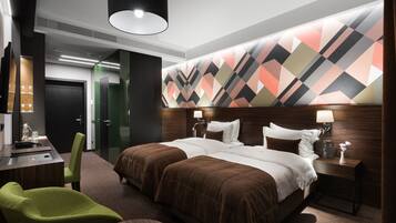 A hotel room with a large bed, two bedside tables, a desk with a chair, a green armchair, and a modern ceiling light.