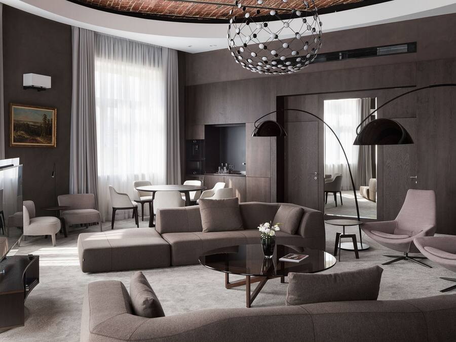 Un salon moderne avec un canapé d’angle, une table basse ronde et un téléviseur. On y trouve également deux fauteuils, une table à manger et un lustre.