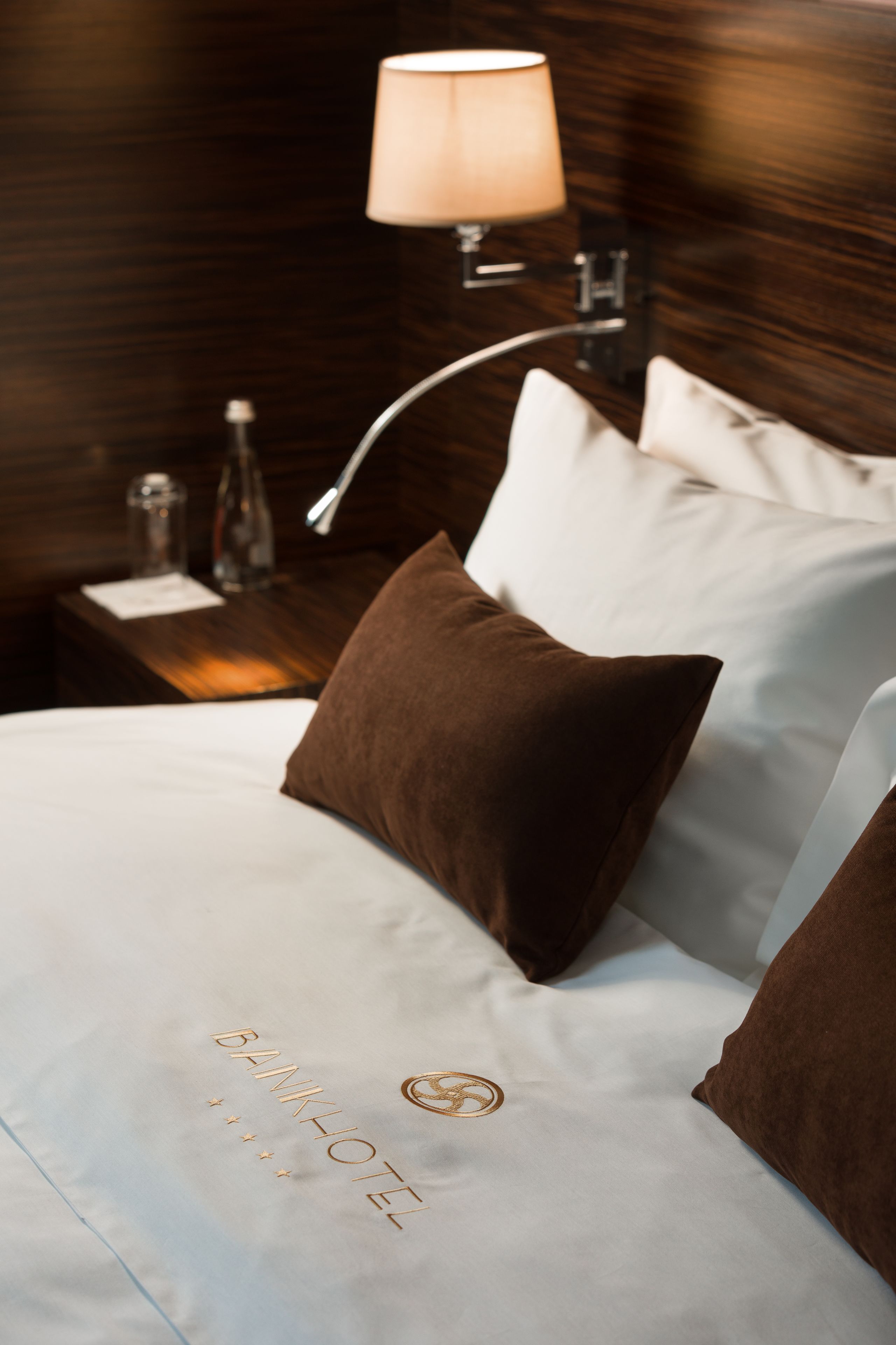 A hotel room with a neatly made bed, a lamp on the nightstand, and a bottle of water.