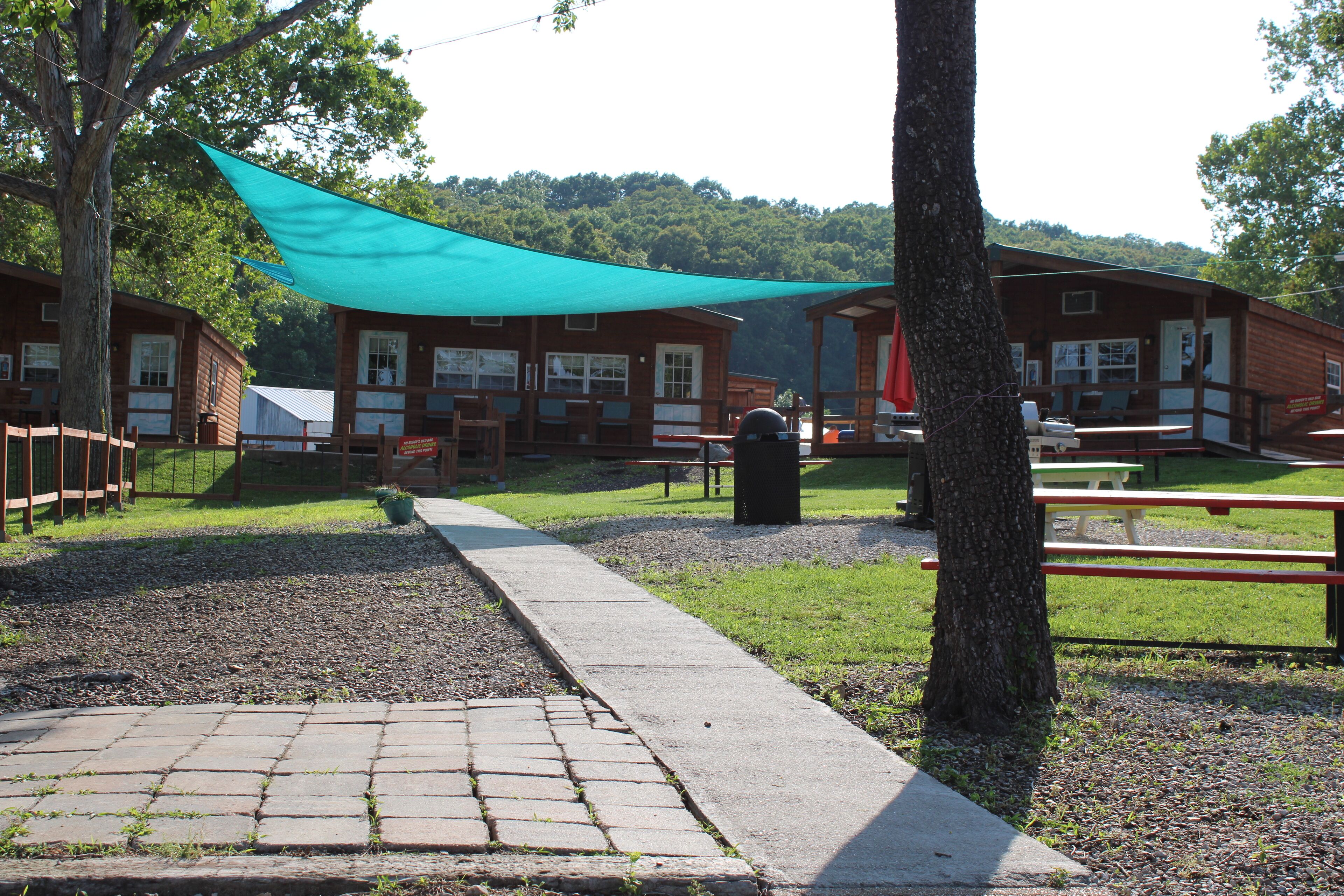 Un camping con cabañas de madera, mesas de picnic y una zona con sombra.