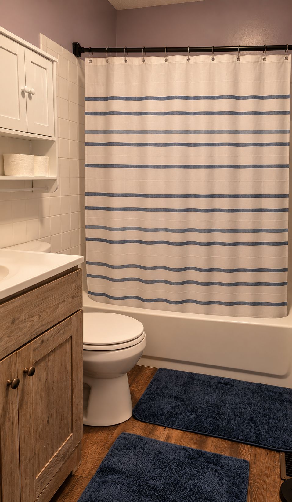 Un baño con inodoro, bañera, un armario de madera, repisa con papel higiénico y una cortina de ducha a rayas.
