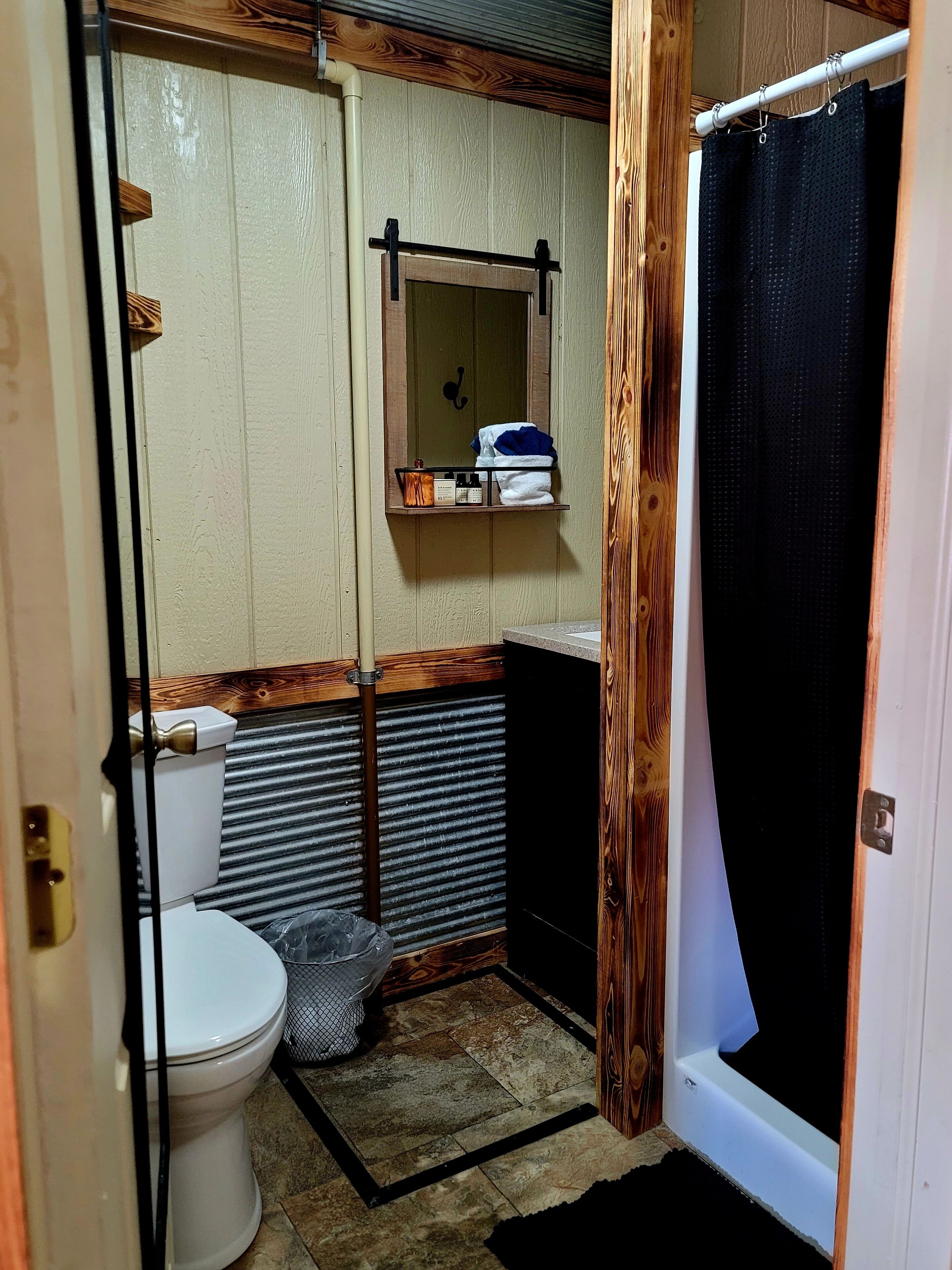 A compact bathroom with a toilet, a small shelf with toiletries, a shower curtain, and a wooden accent wall.