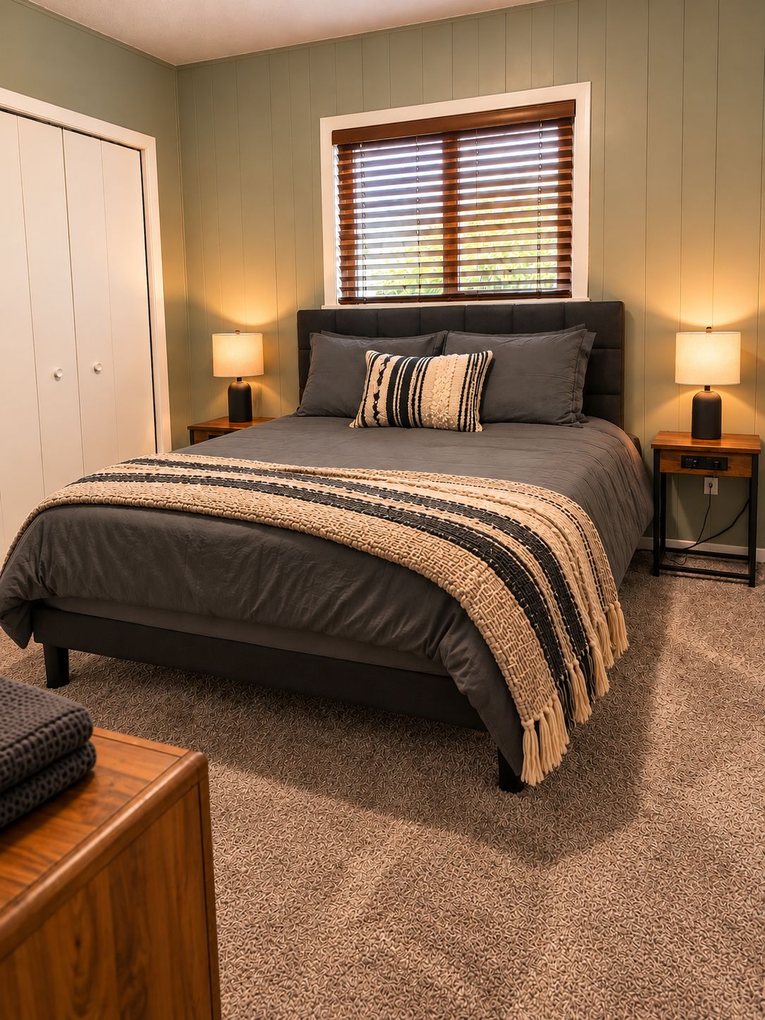 A bedroom with a bed, two bedside lamps, a window with blinds, and a door.