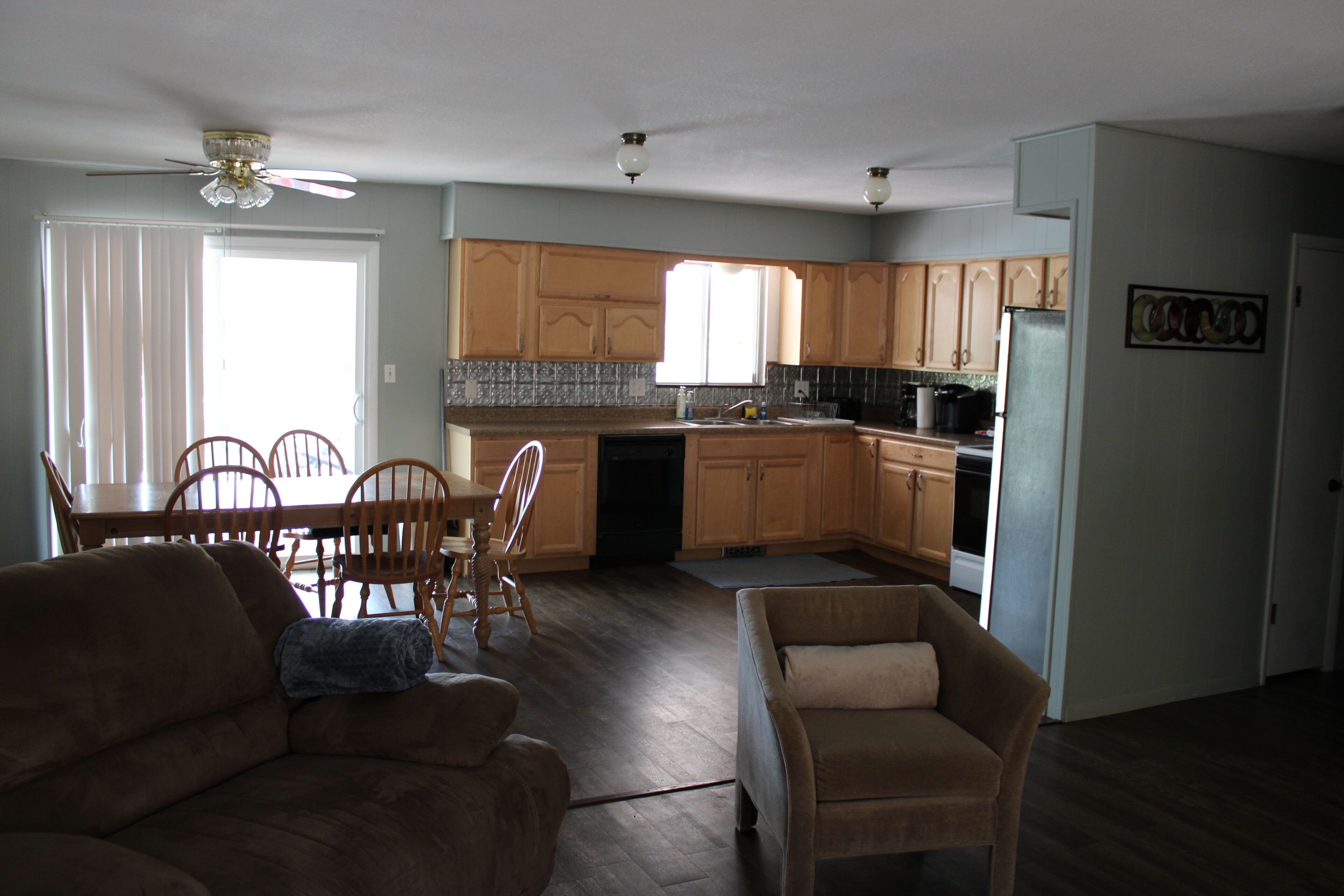 A spacious living room with a brown sofa and a beige armchair, a dining area with wooden chairs and a table, and a kitchen with wooden cabinets and a refrigerator.