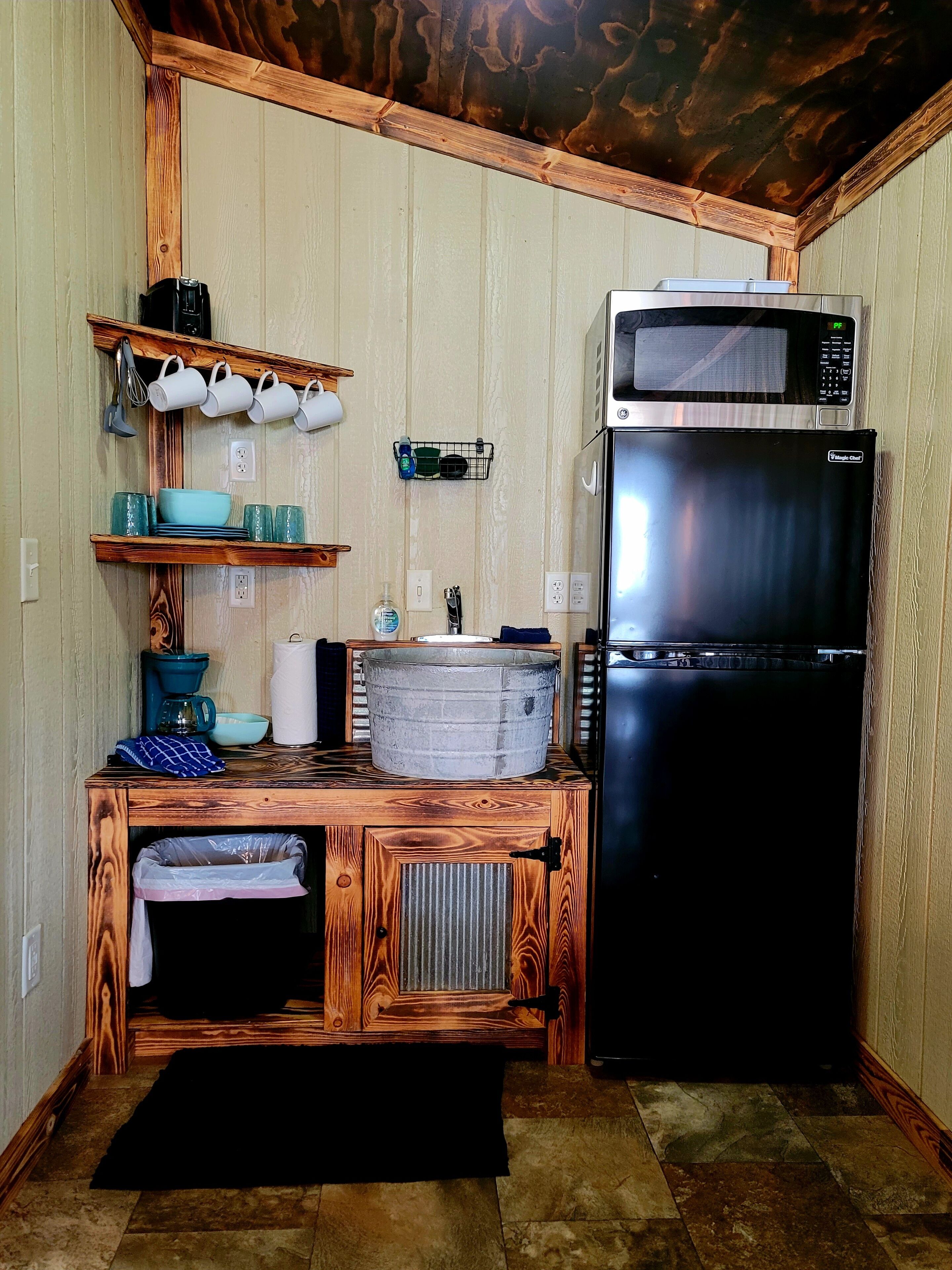 A compact kitchen with a microwave, refrigerator, wooden shelves, and a trash bin.