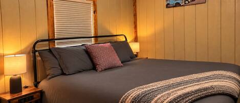 A bedroom with a bed, bedside lamps, a window with blinds, a wall-mounted picture, and a wooden ceiling.
