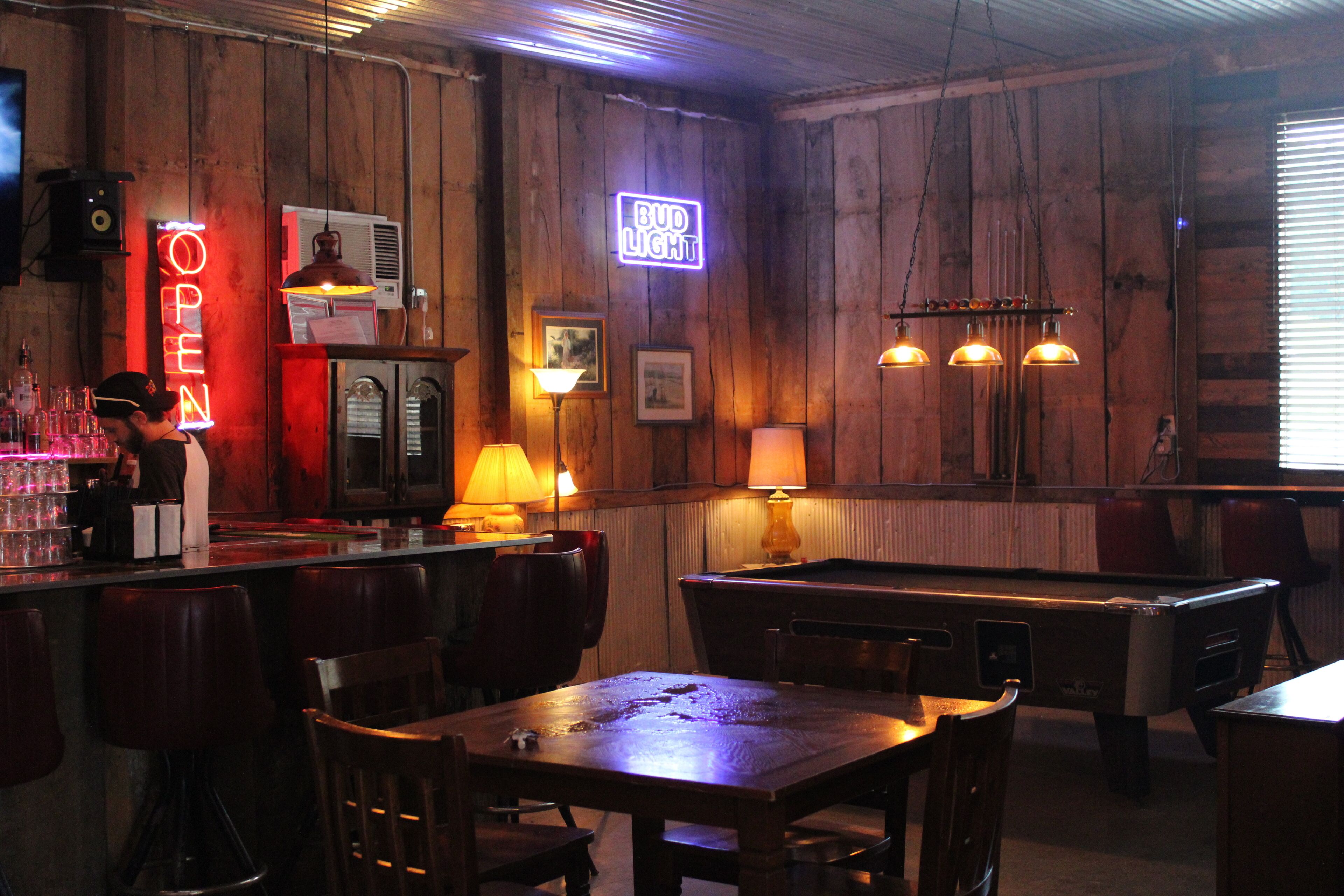 A bar area with wooden walls, a neon 'OPEN' sign, a pool table, and bar stools.