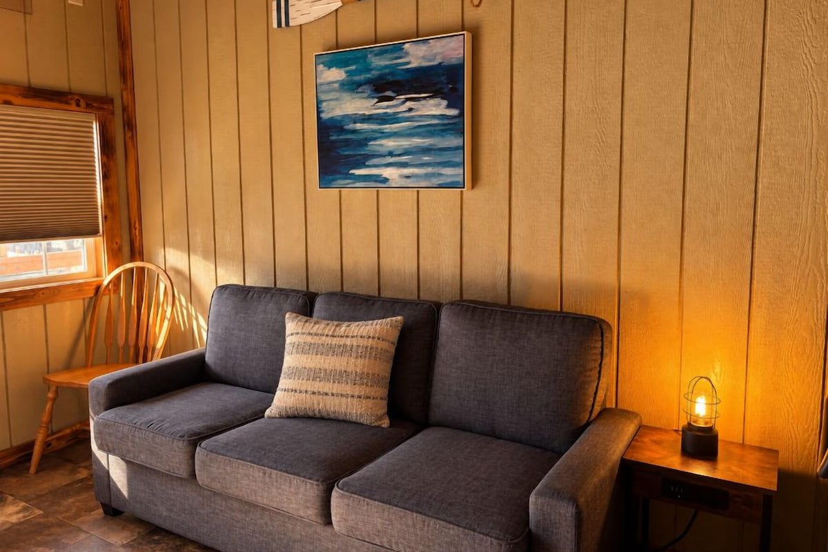 A room with a sofa, a painting, a wooden chair, a lamp, and a window with blinds.