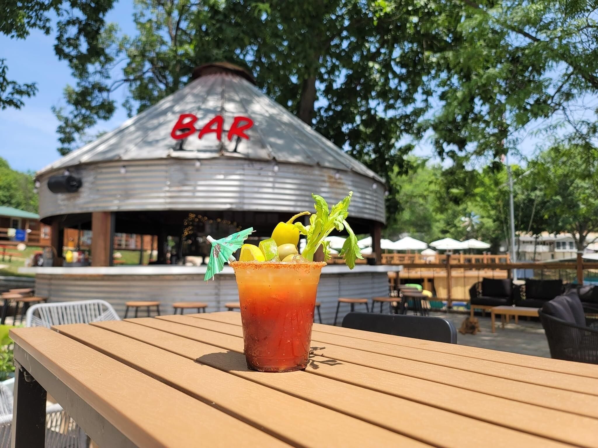 Een houten tafel met een Bloody Mary cocktail, versierd met selderij en een partje limoen. Op de achtergrond een rustieke bar met een bord waarop 'BAR' staat.