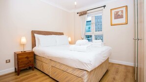 Una cama bien tendida con sábanas blancas y cabecera de madera, una mesita de noche con lámpara y una ventana con cortinas.