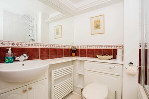 Et badeværelse med hvid vask, toilet, brusebad og spejl. Væggen er beklædt med dekorative fliser, og der hænger et billede i ramme.