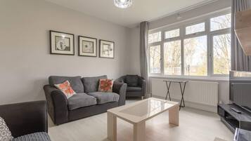 A modern living room with a grey sofa, a coffee table, and a black armchair. There are three framed pictures on the wall, and a window with curtains.