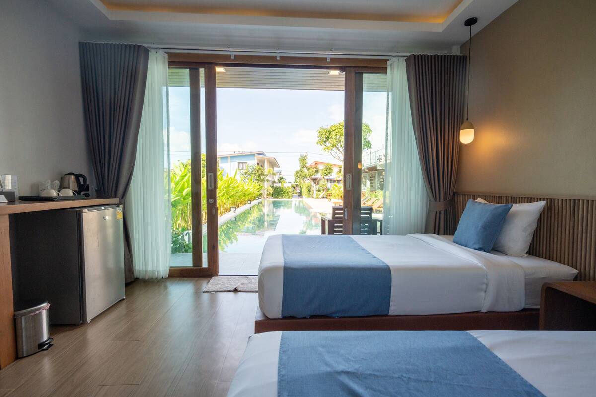 A hotel room with a bed, a desk with a kettle, and a view of a pool and buildings.