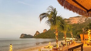 Un espace aménagé avec des tables et des chaises, situé au bord de la plage, doté d’un toit de chaume et offrant une vue sur la mer et les montagnes.
