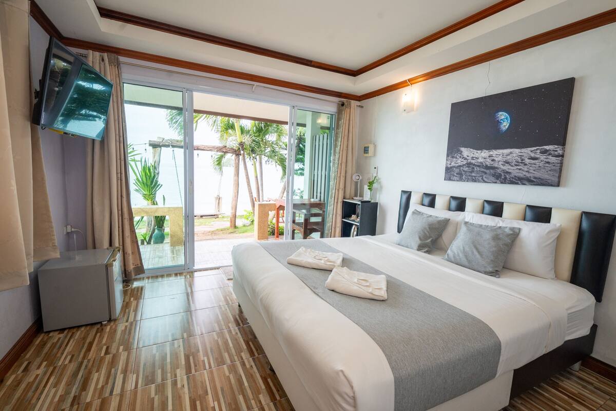A hotel room with a large bed, a flat-screen TV mounted on the wall, and a sliding glass door leading to an outdoor area with palm trees.