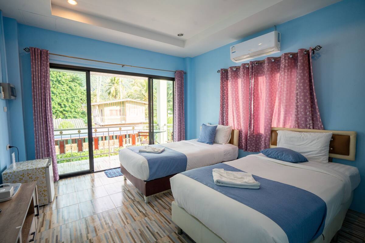 A hotel room with two beds, a balcony with a view of greenery, and an air conditioning unit on the wall.