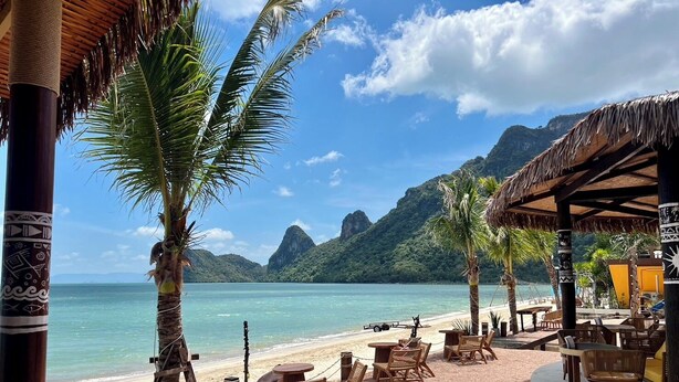 Un restaurant situé en bord de mer, avec une terrasse, des palmiers et une vue sur l’océan et les montagnes.
