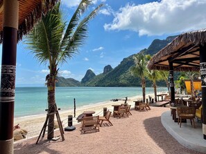 A beachside restaurant with outdoor seating, palm trees, and a view of the ocean and mountains. - Janpa Resort (Don Sak)