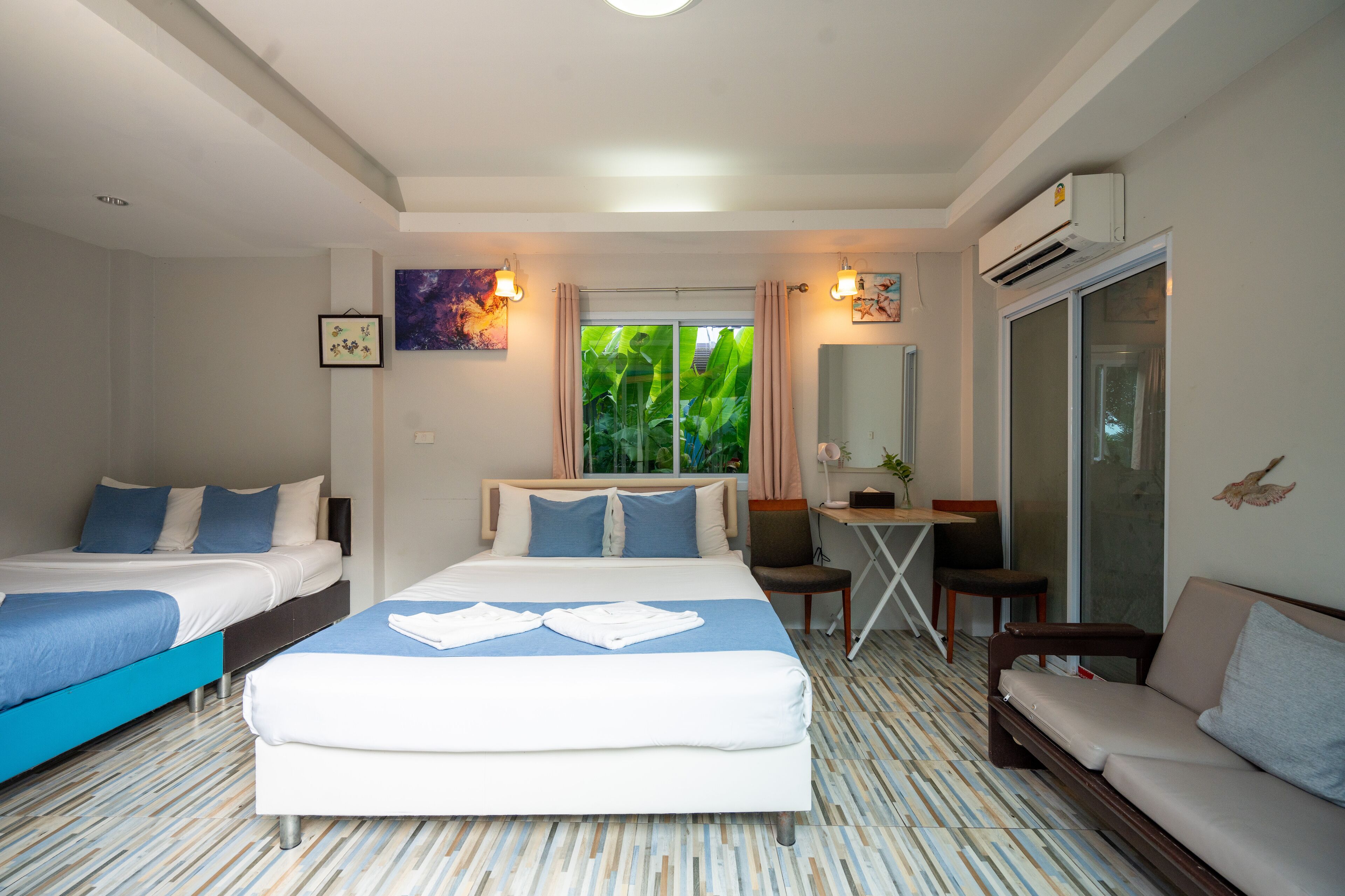 A hotel room with a large bed, a smaller bed, a sofa, a desk, and a chair. There is a window with a view of greenery outside.