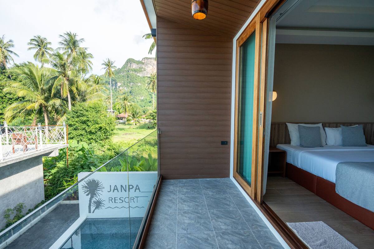 A hotel room with a bed, a balcony with a view of palm trees and a mountain, and a sign that reads "JANPA RESORT".